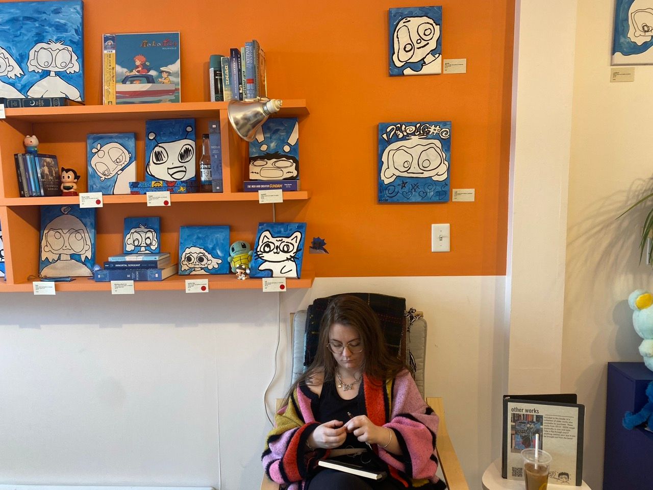 girl sitting in chair with colorful sweater on, blue paintings on orange shelves in background